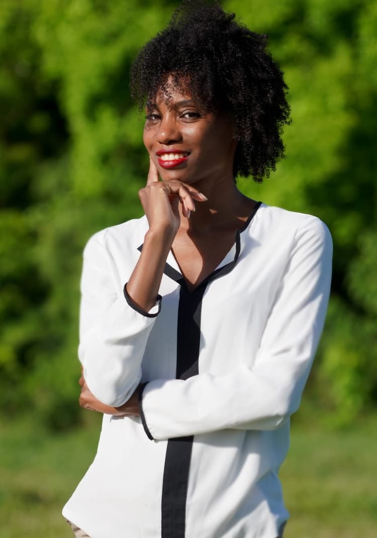 Femme souriante aux cheveux afro naturels portant une chemise blanche avec cravate noire, main posée sous le menton dans une pose confiante sur fond de verdure